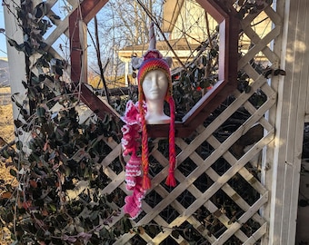 May include: A colorful, handmade crochet unicorn hat with long braids and a ruffled scarf. The hat features a horn and ear details, with a rainbow color scheme. The braids and scarf are shades of pink and red.