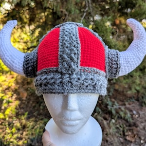 May include: A handmade crochet Viking helmet with white horns, a red crown, and gray trim. The hat is displayed on a white mannequin head against a blurred green background. The horns curve upwards.