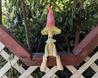May include: A crochet mushroom figure with a tall, pointed cap featuring a gradient of pink, orange, and light green. The body is a pale cream color with a small collar, two black bead eyes, and long, dangling arms and legs in a seated position.