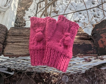 May include: A pair of bright pink knitted fingerless gloves. The gloves have a ribbed cuff and a textured design on the back of the hand, with a small pom-pom detail. The gloves are made of a thick, warm yarn, perfect for cold weather.
