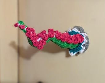 May include: A crocheted wall hanging in the shape of a tentacle. The tentacle is green with pink and white accents, and the base is gray, teal, and white. The handmade piece is likely for home decor.