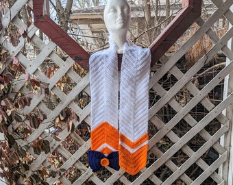 May include: White and orange crocheted scarf with a blue bow. The scarf is hanging on a white lattice fence.