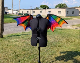 May include: A black dress form displays a pair of crocheted wings. The wings are a vibrant rainbow of colors, with a black border. The wings are attached to the dress form with white accents. The dress form is on a metal stand, outdoors.