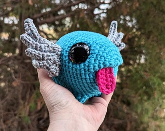 May include: A crocheted blue creature with a pink tongue and a single black eye. The creature has two gray wings.