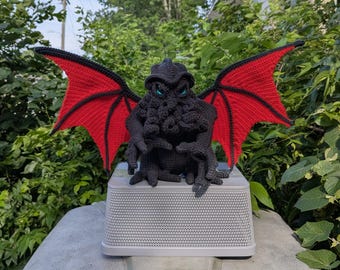 May include: A black crocheted creature with a tentacled face, pointed ears, and large, expansive wings featuring bright red inner panels and black borders. The figure has piercing blue eyes and is crafted with a dense, textured yarn stitch pattern.