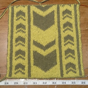 May include: Hand-knitted textile in progress, featuring a geometric arrow pattern in olive green and yellow. The piece is on knitting needles, with a measuring tape showing inches. The design includes vertical stripes of arrows.