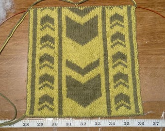 May include: Hand-knitted textile in progress, featuring a geometric arrow pattern in olive green and yellow. The piece is on knitting needles, with a measuring tape showing inches. The design includes vertical stripes of arrows.