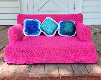 May include: A pink crocheted couch with three blue and green crocheted pillows. The couch is sitting on a wooden floor.