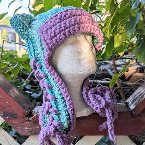 May include: A crocheted hat with a purple and teal color gradient. The hat has two ear flaps and a long, flowing scarf attached to the back.
