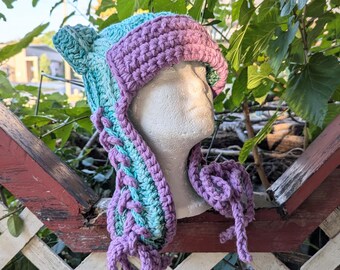 May include: A crocheted hat with a purple and teal color gradient. The hat has two ear flaps and a long, flowing scarf attached to the back.