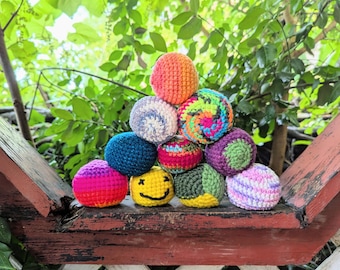 May include: A pyramid stack of ten small, round, crocheted yarn balls in various colors and patterns, including a yellow ball with a black stitched smiley face, solid bright pink, orange, blue, purple, green, and multi-colored variegated yarn.