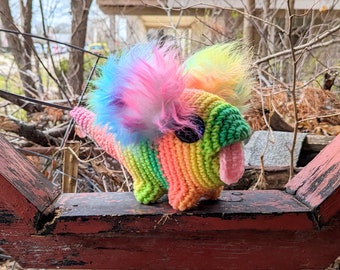 May include: A rainbow crocheted dinosaur toy with a pink tongue and a fluffy, multi-colored mane. The toy is sitting on a wooden fence.