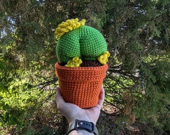 May include: A handmade crochet cactus in a terracotta-colored pot. The cactus is green with yellow accents and sits in a brown base. The pot is a vibrant orange color. The item is held in a person's hand against a backdrop of green foliage.