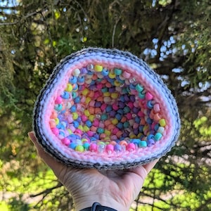 May include: A crocheted geode-shaped container, filled with colorful beads. The geode has a gray and white exterior, a pink interior, and is held in a hand against a backdrop of green foliage. The beads are various colors.