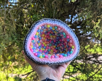 May include: A crocheted geode-shaped container, filled with colorful beads. The geode has a gray and white exterior, a pink interior, and is held in a hand against a backdrop of green foliage. The beads are various colors.