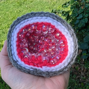 May include: A crocheted geode-shaped item with a gray, white, and red exterior. The center is filled with clear, iridescent beads. The geode is held in a hand against a backdrop of green grass and foliage.