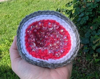 May include: A crocheted geode-shaped item with a gray, white, and red exterior. The center is filled with clear, iridescent beads. The geode is held in a hand against a backdrop of green grass and foliage.