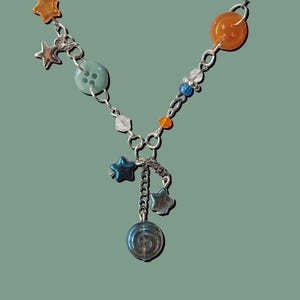 May include: A silver-toned necklace with a variety of colorful beads and charms. The necklace features orange, blue, and clear beads, along with star-shaped and button-shaped charms. A spiral charm hangs from the center.