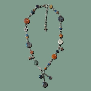May include: A silver-toned necklace with a celestial theme. It features orange, blue, and silver beads, star charms, and a dangling pendant. The necklace is displayed against a muted green background.