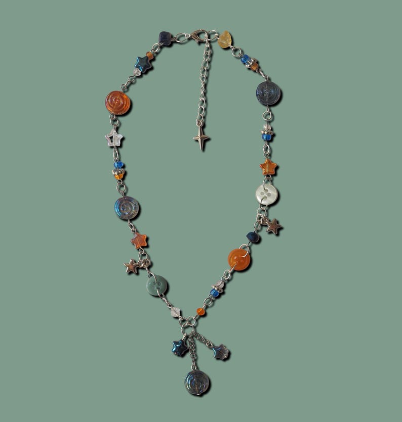 May include: A silver-toned necklace with a celestial theme. It features orange, blue, and silver beads, star charms, and a dangling pendant. The necklace is displayed against a muted green background.