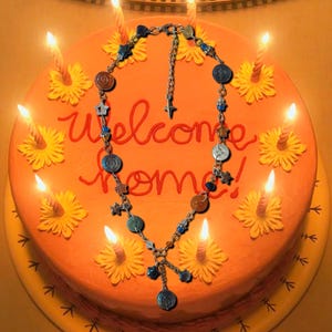 May include: A necklace with various charms, including stars and circles, is displayed on an orange cake decorated with yellow flowers and lit candles. The cake has the words "Welcome Home" written in red icing.