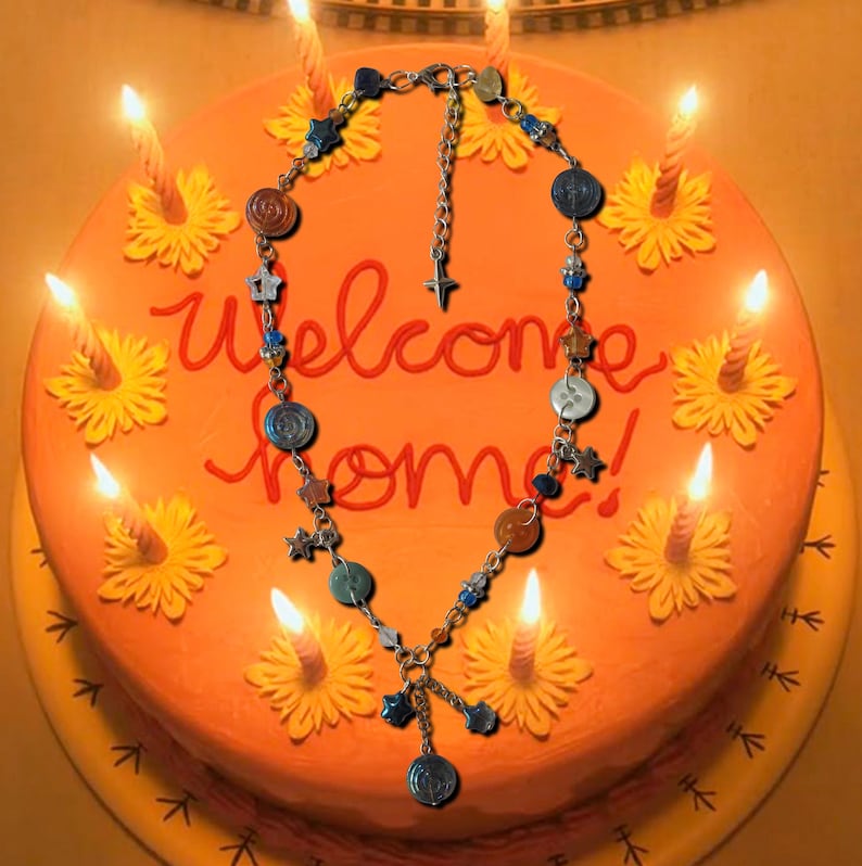 May include: A necklace with various charms, including stars and circles, is displayed on an orange cake decorated with yellow flowers and lit candles. The cake has the words "Welcome Home" written in red icing.