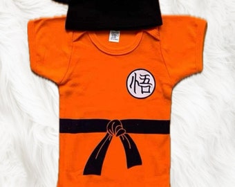 May include: Orange baby onesie with a black belt and a white circle with the kanji for "悟" (悟) on the front. The onesie is paired with a black hat with two points on top.