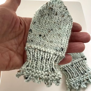 May include: A pair of knitted baby mittens in a light blue color with flecks of brown and black. The mittens have a ruffled cuff design.