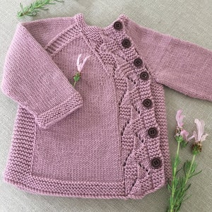 Op de afbeelding: Een mauve gebreide babytrui met knoopsluiting en een kabelbreipatroon.