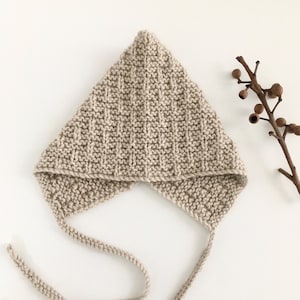 May include: A beige knitted baby bonnet with a long tie. The bonnet is made with a simple stitch pattern and has a pointed shape.