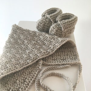 Puede incluir: Un conjunto de gorro y botines de punto para bebé en color beige claro. El gorro tiene una larga cinta que se puede atar debajo de la barbilla. Los botines tienen un diseño sencillo con la punta puntiaguda.