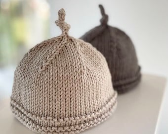 May include: Two knitted hats, one beige and one brown, with a unique knot detail on top. The hats are made with a textured stitch and have a folded brim.