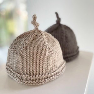 May include: Two knitted hats, one beige and one brown, with a unique knot detail on top. The hats are made with a textured stitch and have a folded brim.