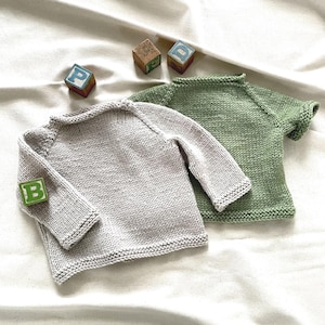 May include: Two hand-knitted sweaters, one in a light beige color with long sleeves and one in a sage green color with short sleeves. Both sweaters feature a simple stockinette stitch pattern with garter stitch edges at the neck, cuffs, and hem.