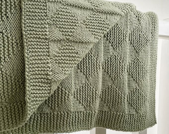 May include: A sage green knitted throw blanket with a geometric pattern. The blanket is folded and draped over a white railing.