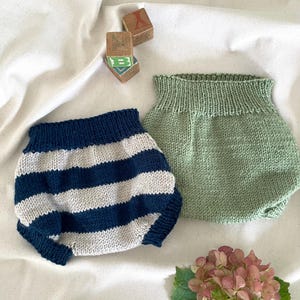Puede incluir: Dos bloomers de bebé tejidos. Uno es de rayas azul marino y blancas, el otro es de color verde claro liso. Bloques de letras de madera están en el fondo. Una flor de hortensia rosa está en primer plano.