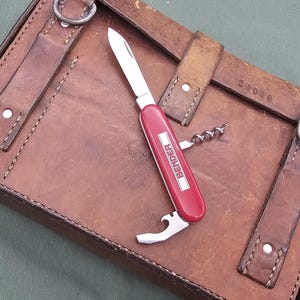 May include: A red multi-tool knife with the word "BENDER" printed on the side, resting on a brown leather case. The knife has a blade, corkscrew, and bottle opener. The case has straps and metal rings.