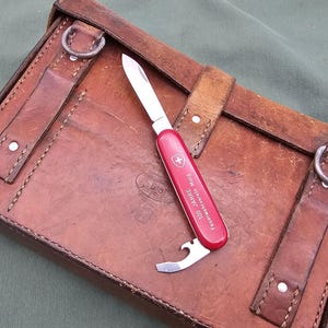 May include: A red and silver Swiss Army knife with a bottle opener and a blade, resting on a brown leather case. The knife has text and a white cross logo. The case has straps and metal rings.