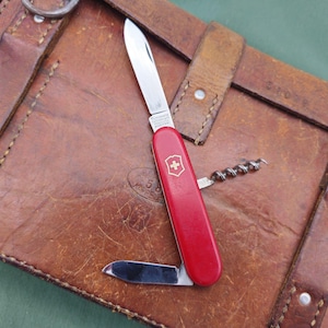 May include: A red Swiss Army knife with several stainless steel blades and a corkscrew, placed on a brown leather surface. The knife displays the Swiss cross emblem. The leather surface has straps and metal rings.