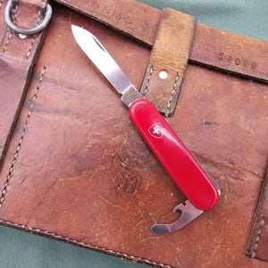 May include: A red Swiss Army knife with a silver blade and bottle opener, featuring the Swiss cross emblem. The knife rests on a worn, brown leather case with straps and metal hardware, suggesting a vintage or utilitarian theme.