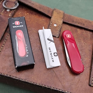May include: A red Wenger Swiss Army knife with a white cross emblem, alongside its black box and instruction manual. The knife is shown open and closed. The items rest on a brown leather surface.
