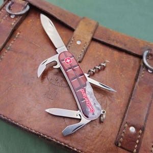 May include: A Swiss Army knife with a red and brown chocolate bar design, featuring multiple tools including a blade, bottle opener, and corkscrew. The knife is set on a brown leather case with metal rings and straps. The text "Switzerland!" is visible.