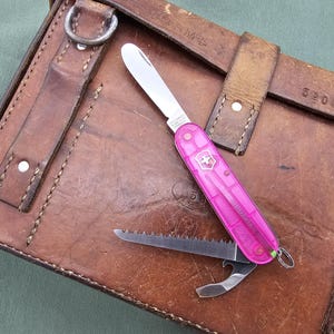 May include: A pink Swiss Army knife with multiple tools, including a blade and saw, rests on a worn, brown leather satchel. The knife features the iconic Swiss cross emblem. The satchel has visible stitching and metal hardware.