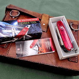 Könnte beinhalten: Ein rotes Victorinox Cyber Tool Multi-Tool in einer weißen Box, mit einer Bedienungsanleitung und Produktverpackung. Die Artikel befinden sich auf einer braunen Ledertasche. Die Verpackung trägt den Text "Cyber Tool".