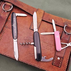 May include: An assortment of pocket knives and a pink Swiss Army knife rest on a brown leather satchel. The knives have black and silver blades, with one featuring a corkscrew and bottle opener. The pink knife has a cross emblem.