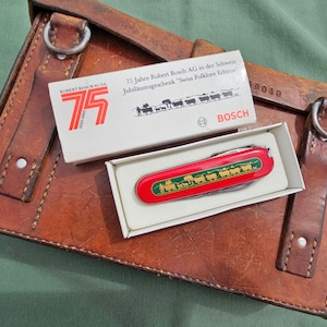 Könnte beinhalten: Ein rotes Multifunktions-Taschenmesser mit grün-goldener Kuh-Grafik in einer weißen Schachtel mit der Aufschrift 75 Jahre Robert Bosch AG in der Schweiz Jubiläumsgeschenk Swiss Folklore Edition, liegend auf einer braunen Ledertasche.