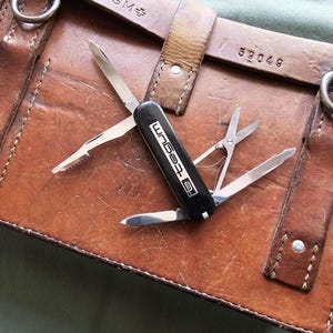 Op de afbeelding: Een bruine leren tas met een zwart multi-tool met het woord "Tegum" in het wit. De multi-tool heeft een mes, schaar en andere gereedschappen. De tas heeft een riem en metalen ringen.