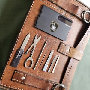 Könnte beinhalten: Ein brauner Leder-Organizer mit verschiedenen Werkzeugen, darunter ein kleines Messer, eine Schere und eine Nagelfeile. Ein schwarzer Kartenhalter mit Logo ist ebenfalls vorhanden. Der Organizer hat Riemen und Metallringe.