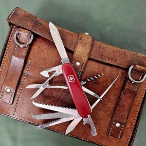 Könnte beinhalten: Ein rotes Schweizer Taschenmesser mit mehreren Werkzeugen, darunter eine Klinge, eine Säge und ein Flaschenöffner, wird geöffnet auf einer braunen Ledertasche gezeigt. Die Tasche hat Metallringe und Riemen. Der Hintergrund ist ein gedämpftes Grün.