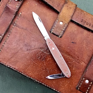 Könnte beinhalten: Ein rotes und silbernes Schweizer Taschenmesser mit ausgeklappter kleiner Klinge, das auf einem abgenutzten braunen Lederetui liegt. Das Messer hat ein Kreuzemblem und einen strukturierten Griff. Das Etui hat Lederriemen und Metallnieten.